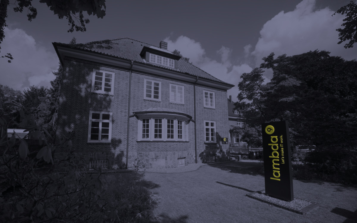 Front view of a villa surrounded by greenery. Next to the villa stands an illuminated sign reading ‘lambda9’.